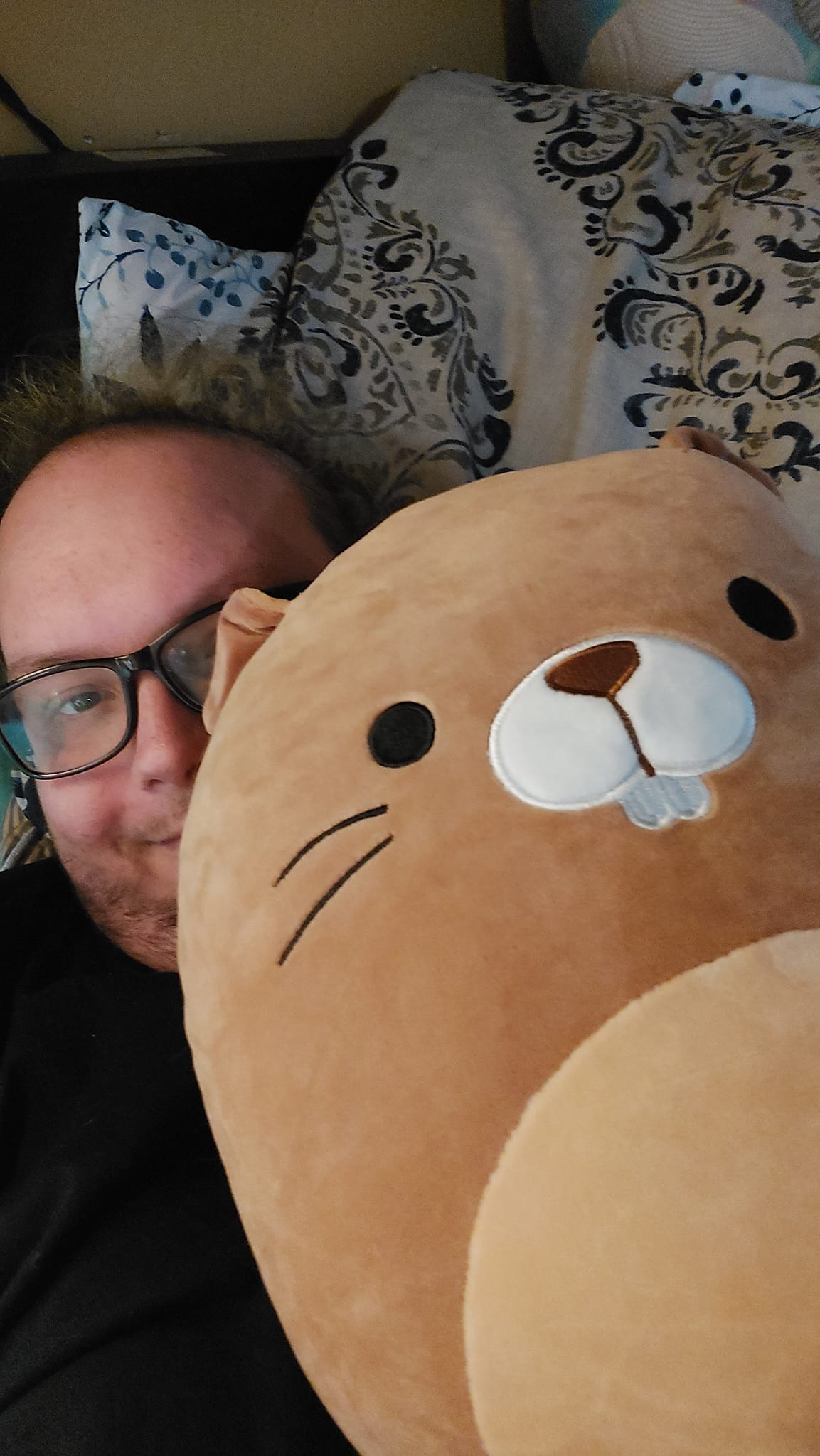 A man with thin green hair lies in bed, holding a beaver Squishmallow